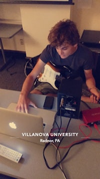 a young man sitting at a desk with a laptop and a guitar